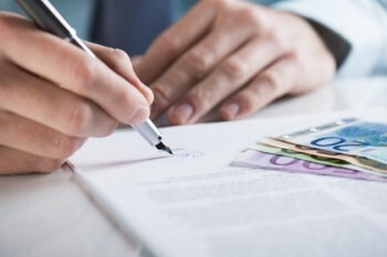 Eine Hand unterzeichnet einen Vertrag, auf dem Vertrag liegen unterschiedliche Geldscheine