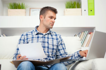 Gesundheitsmanagement Master Fernstudent sitzt bei sich auf dem Sofa und lernt mit Unterlagen und Laptop