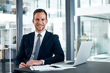 Betriebswirt sitzt im Anzug am Schreibtisch mit Laptop.