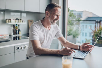 Mann, der ein Sicherheitsmanagement Fernstudium absolviert, sitzt in der Küche am Laptop und lernt