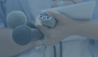 Ein/e Journalist/in hat zwei Mikrofone, einen Notizblock und ein Aufnahmegerät in den Händen.