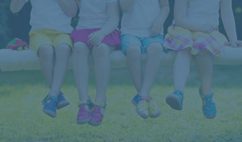 Vier Kinder sitzen auf einer Bank, haben bunter Sommerschuhe an und lassen die Füße baumeln