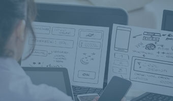 Eine Medieninformatikern sitzt vor verschiedenen Bildschirmen und einer Interface-Skizze.