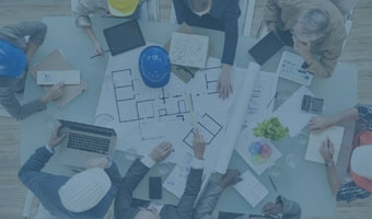 Vogelperspektive auf einen Schreibtisch an dem acht Ingenieure sitzen, die an einem Bauplan arbeiten.